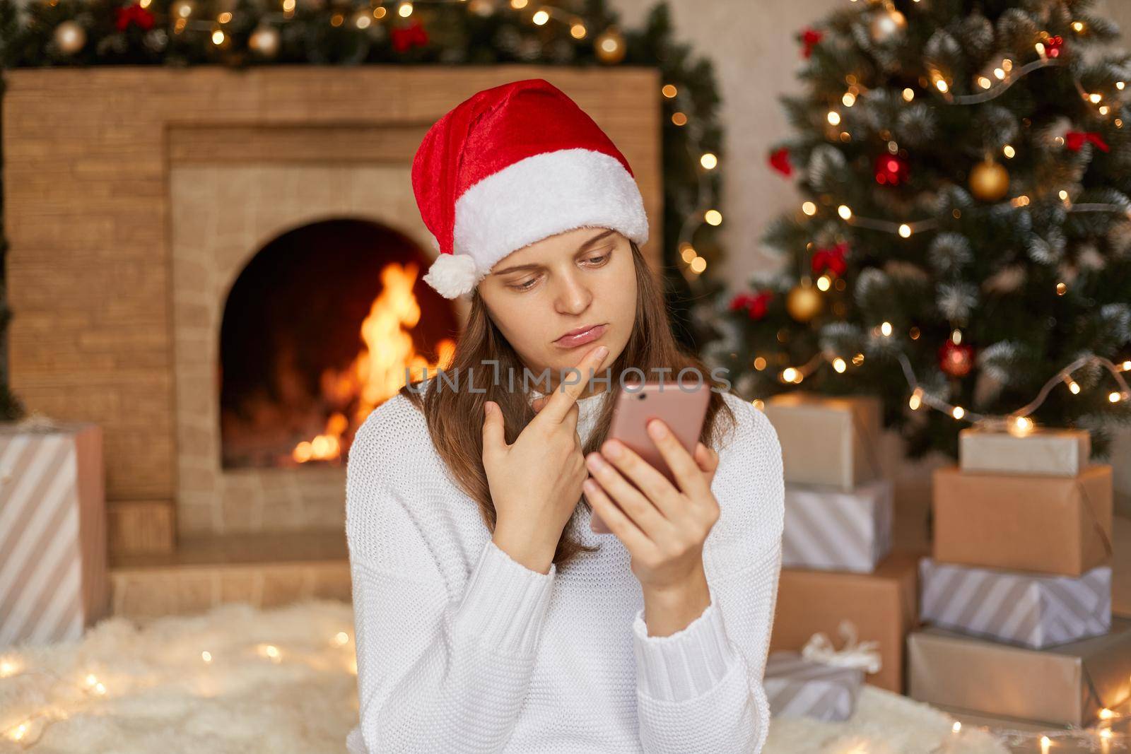 Upset female wearing white sweater and santa hat chatting with somebody. woman reads sms on phone, being in bed mood, keeping finger on chin, looks at device's screen with sad expression.