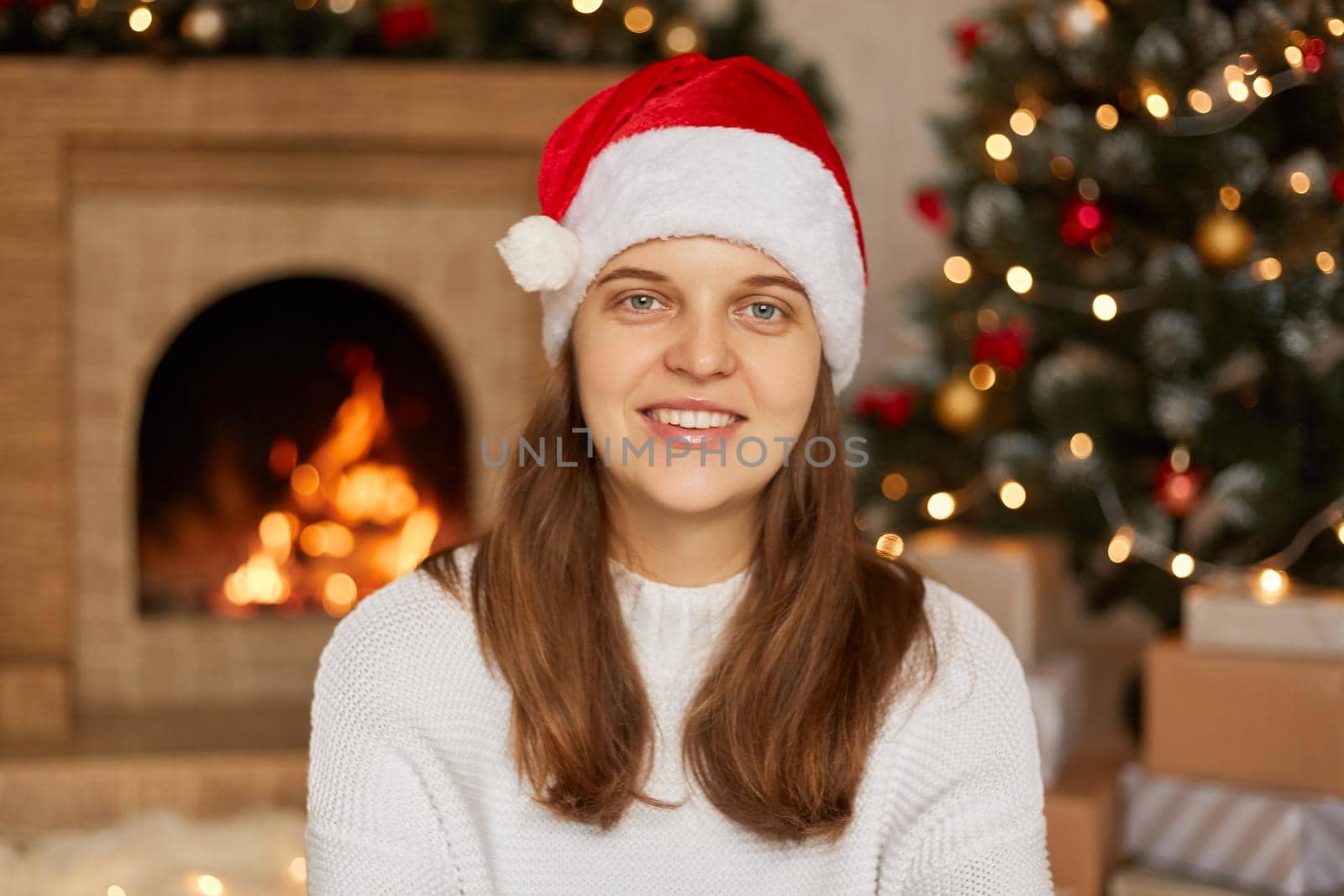 Portrait of beautiful Caucasian female model wear santa hat and white sweater posing at home with fireplace and x-mas tree on background, lady with toothy smile looks at camera.
