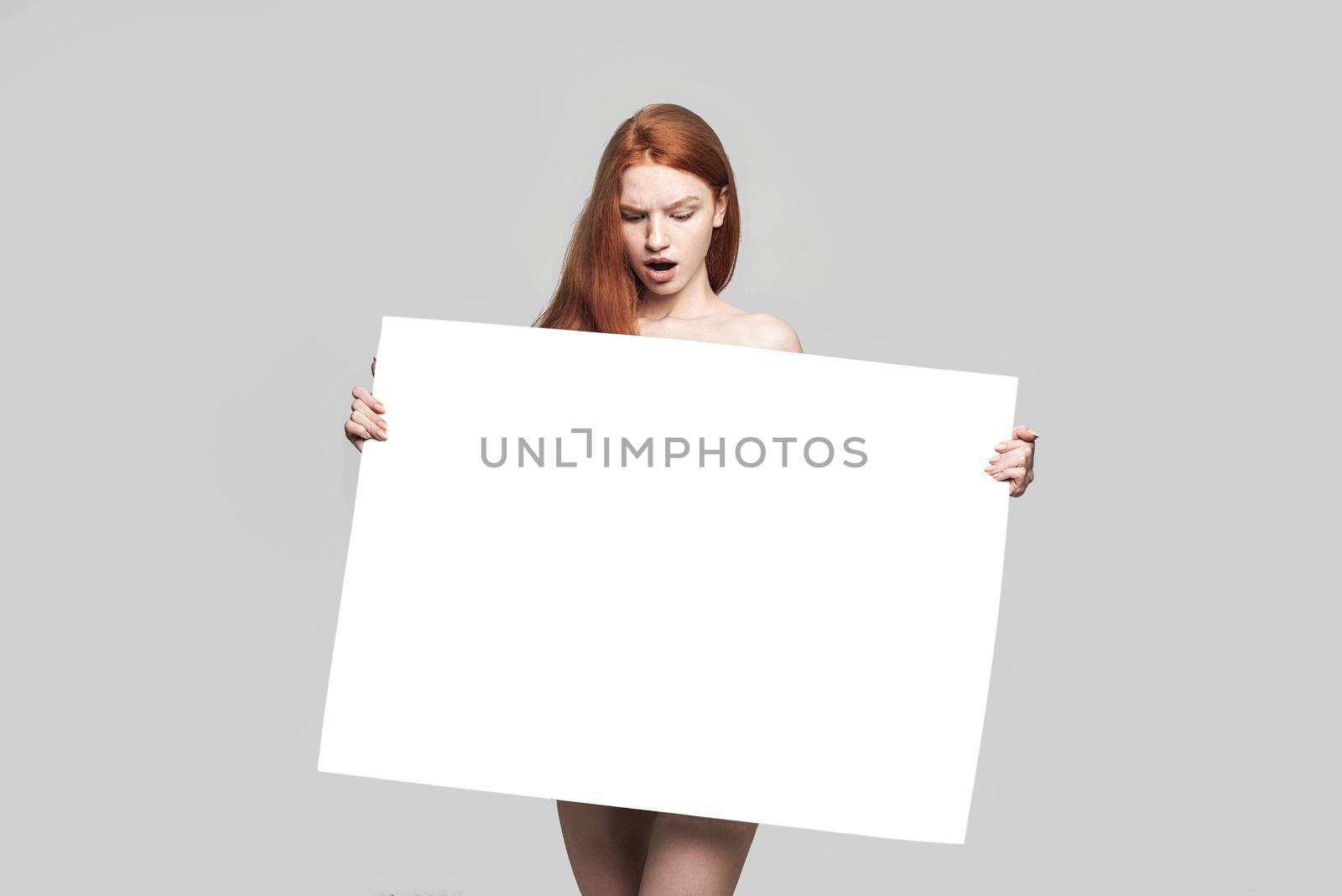 Studio shot of shocked young redhead woman holding empty blank board and looking at it while standing against grey background. Advertising