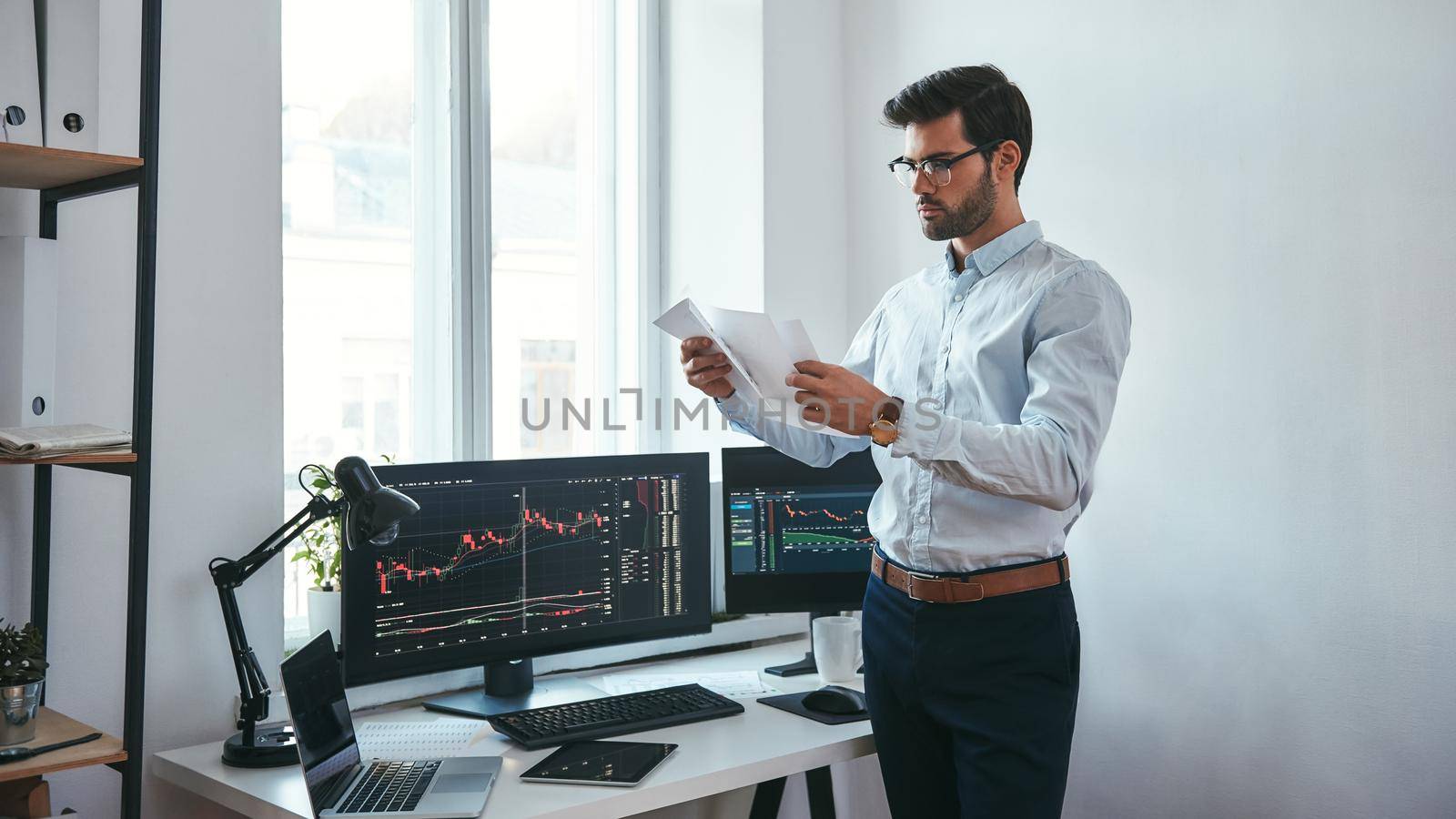 Analyzing data. Thoughtful trader in eyeglasses holding a financial report and analyzing trading charts while standing in front of computer screens in modern office. Stock broker. Forex market. Trade concept