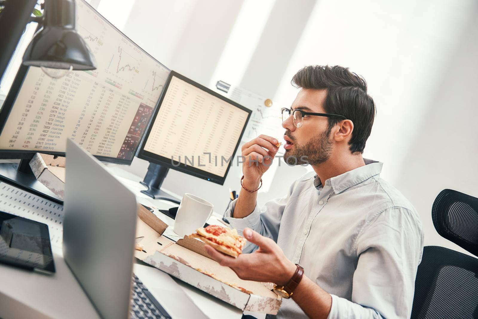 It can not be truth Disappointed young trader is looking at monitor screen with trading charts and financial data while eating hot pizza in his modern office. Food concept. Business concept. Trade concept. Bad news