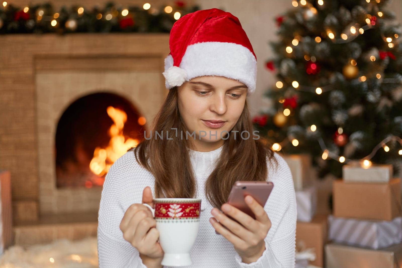 Beautiful young woman drinking tea, wearing santa hat and sweater, using her mobile phone in christmas, reading news from social networks, looks calm and concentrated, poses in decorated living room.
