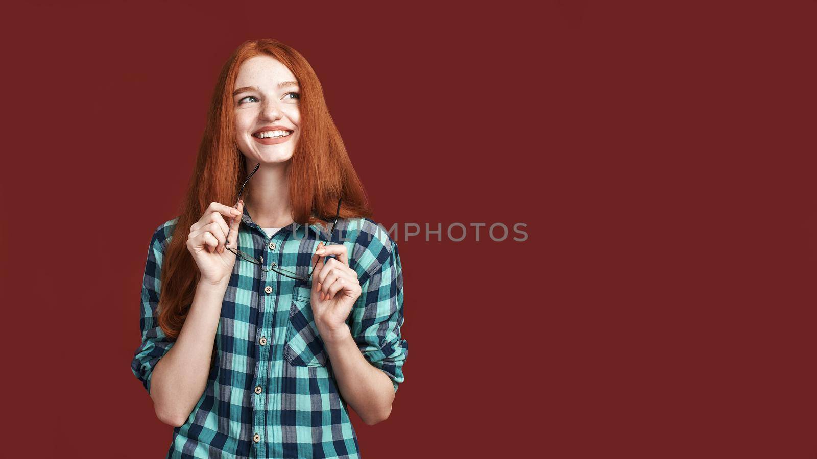 Glad, joyful, carefree girl with beautiful red long hair holding glasses and laughing while standing over red background