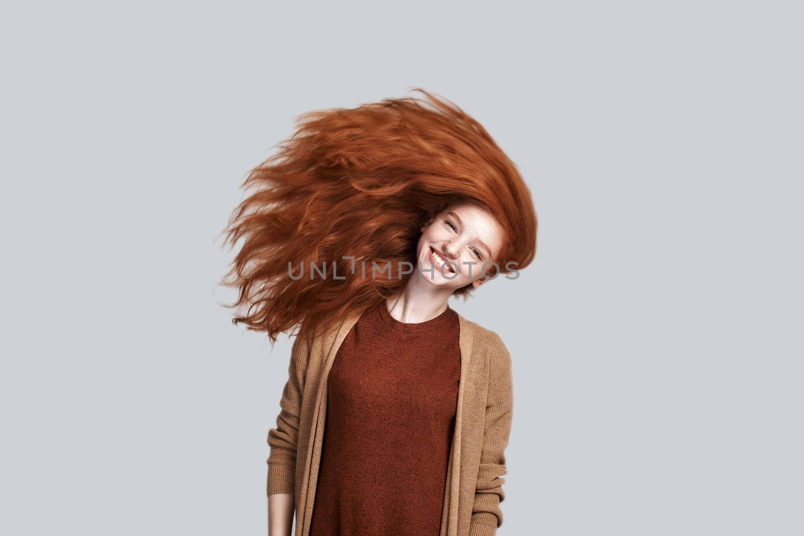 Studio shot of cheerful and young redhead woman with tousled hair smiling at camera while standing against grey background Beauty concept. Happiness
