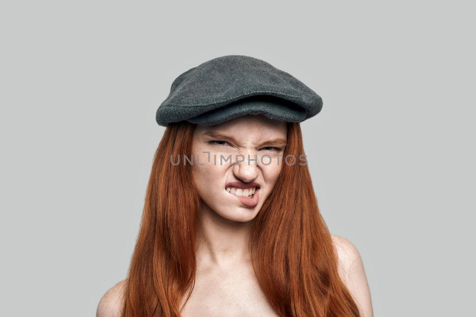 Rude girl. Portrait of young dissatisfied redhead woman in headgear biting a lip and looking at camera while standing against grey background by friendsstock
