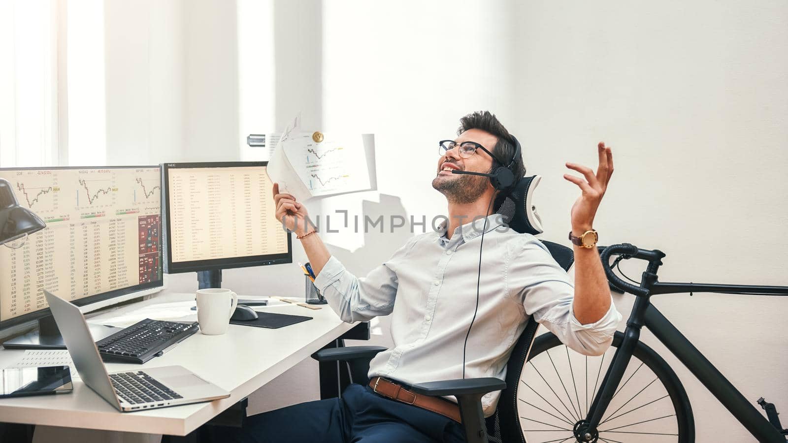 Great Happy and successful bearded trader in headset holding financial report, talking with client and gesturing while sitting in front of monitor screens in the office. Success concept. Trade concept. Call center