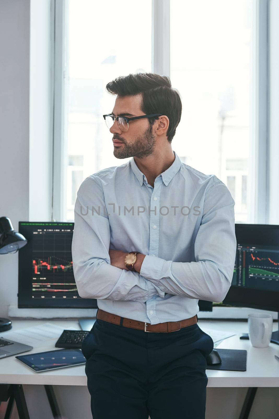 Young and smart. Vertical photo of confident businessman or trader in formal clothes and eyeglasses looking away and keeping arms crossed while standing at office. by friendsstock