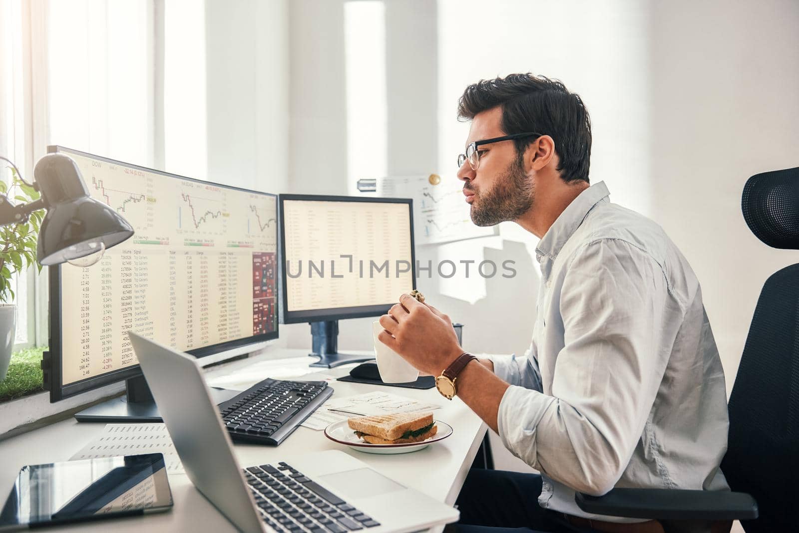 Hungry businessman. Side view of young bearded trader is eating fresh sandwich while looking at monitor screen with financial data in his modern office by friendsstock