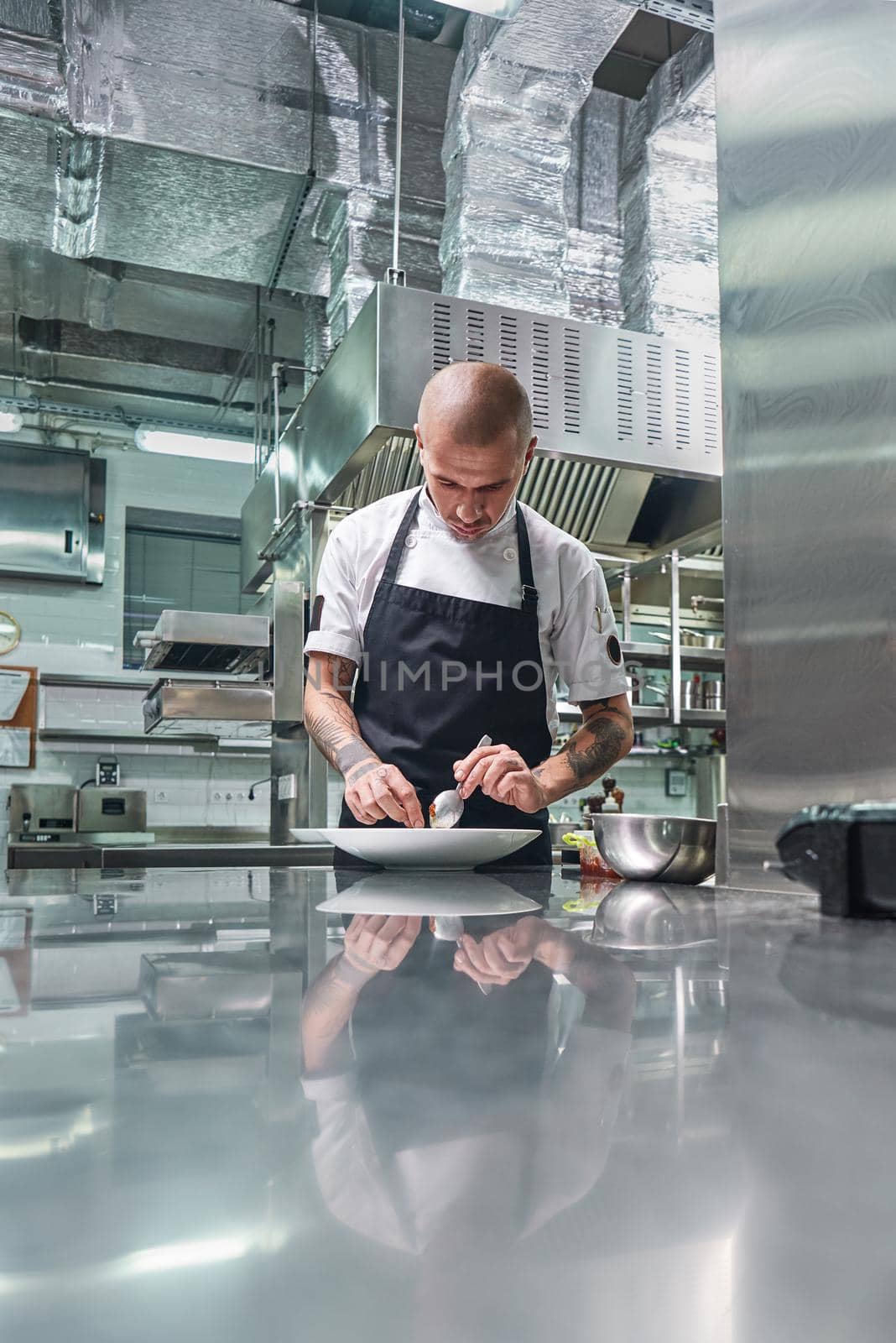 Working in a restaurant. Vertical portrait of professional male chef with tattoos on his arms garnishing his dish on the white plate while standing in a kitchen