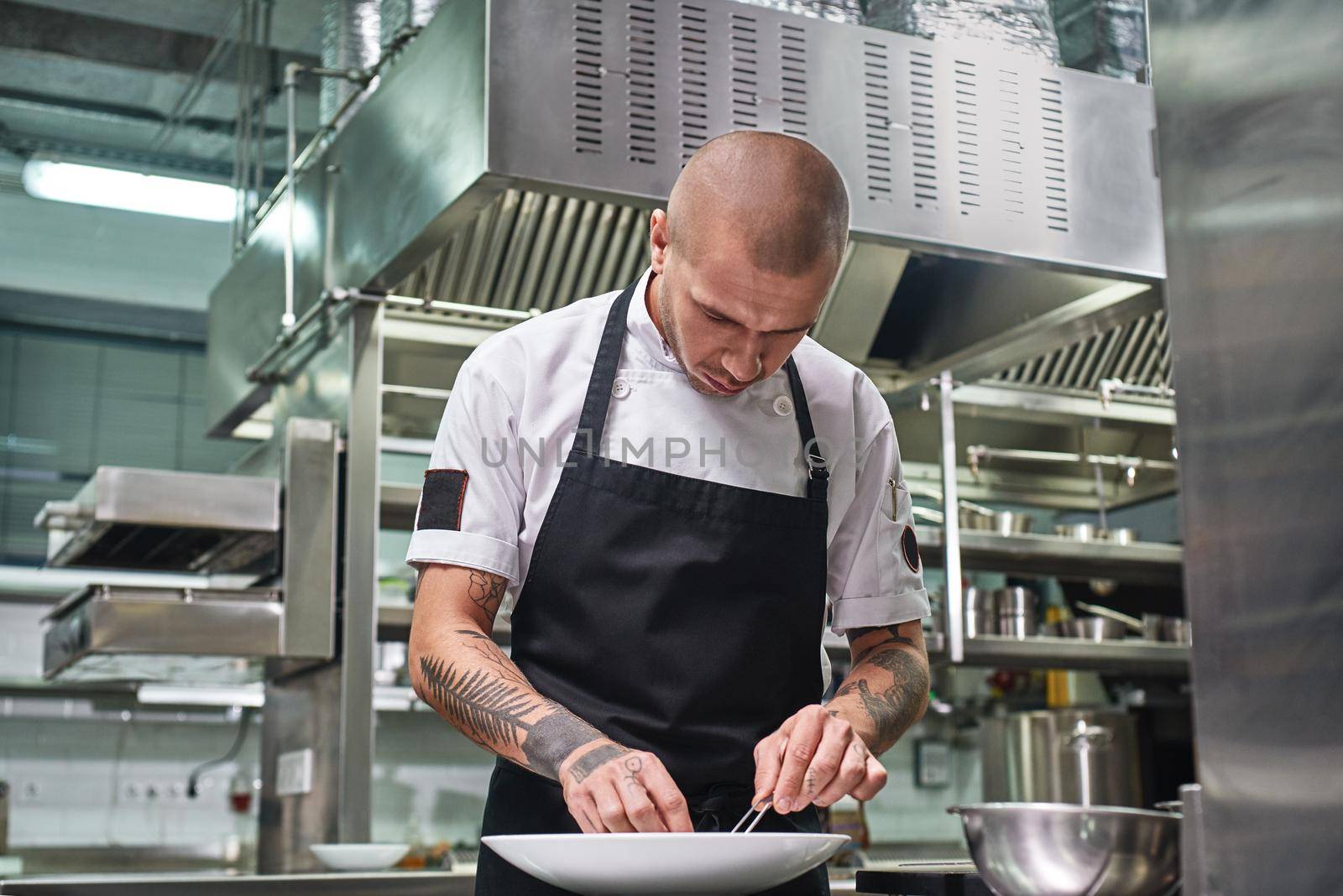 Finishing a dish. Attractive male chef with beautiful tattoos on his arms garnishing his dish on the plate in restaurant kitchen. Food decoration