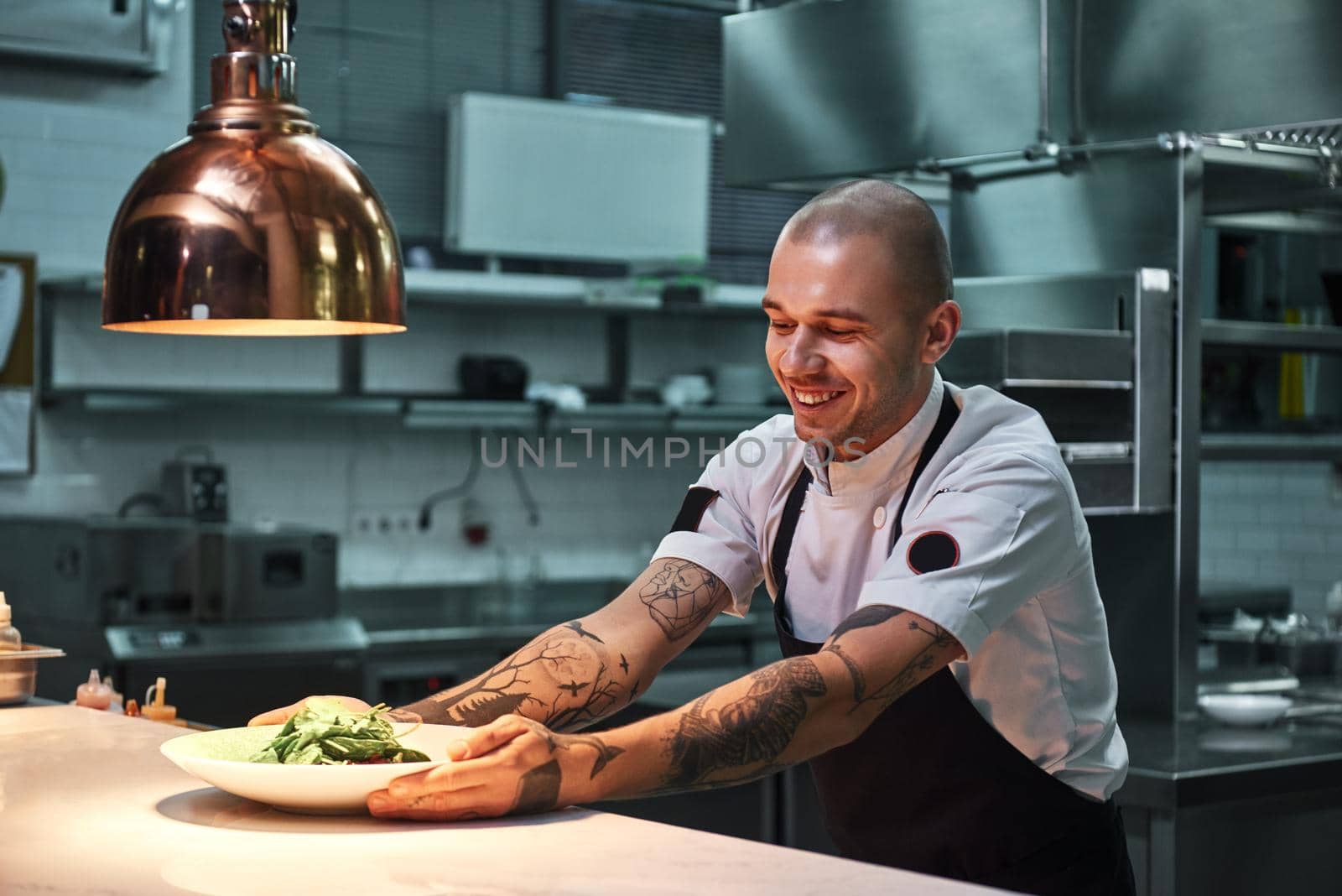 Ready to serve. Young smiling chef with tattoos on his arms, in black apron holding ready dish in modern restaurant kitchen. Food concept