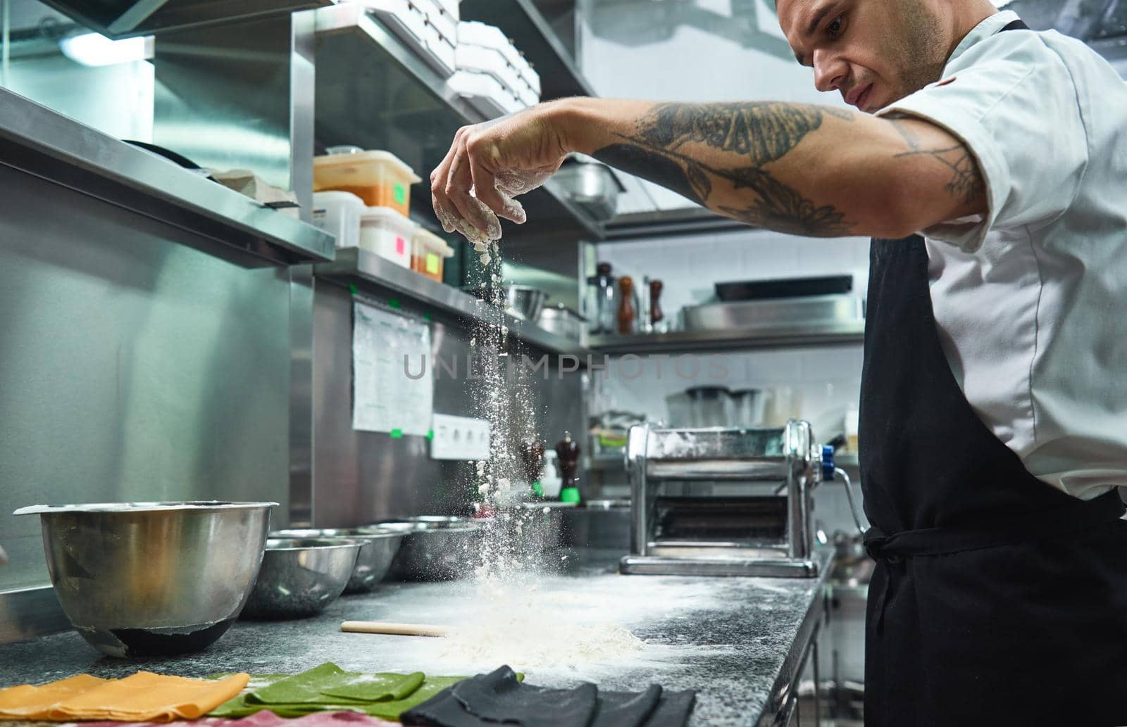 Slow motion. Handsome young chef with black tattoos on his arms pouring flour on kitchen table before making pasta by friendsstock