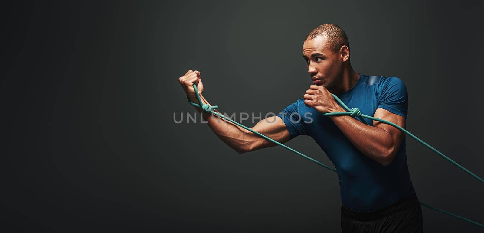 Isolated studio portrait of clean shaven young dark skinned male athlete with strong muscular shoulders working out using resistance elastic band, strengthening arm muscles over dark background. Side view