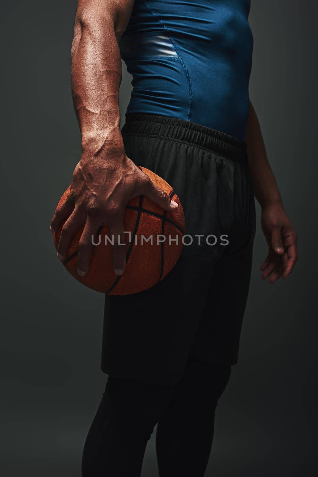 Close up of basketball player in blue T-shirt and black shorts standing isolated over dark background and holding a ball