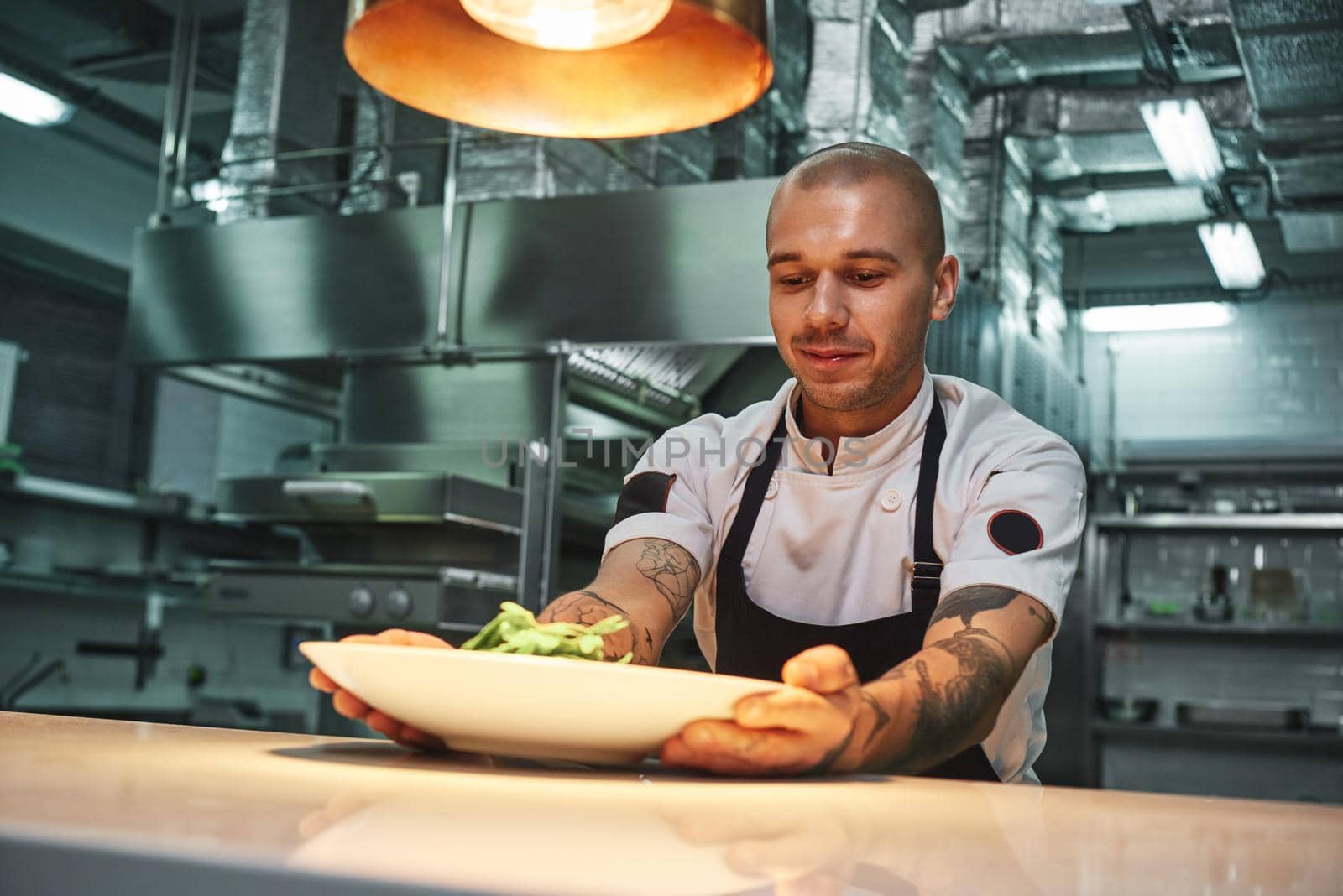 Ready to eat. Handsome smiling chef with tattoos on his arms, in black apron holding ready dish in modern restaurant kitchen. by friendsstock