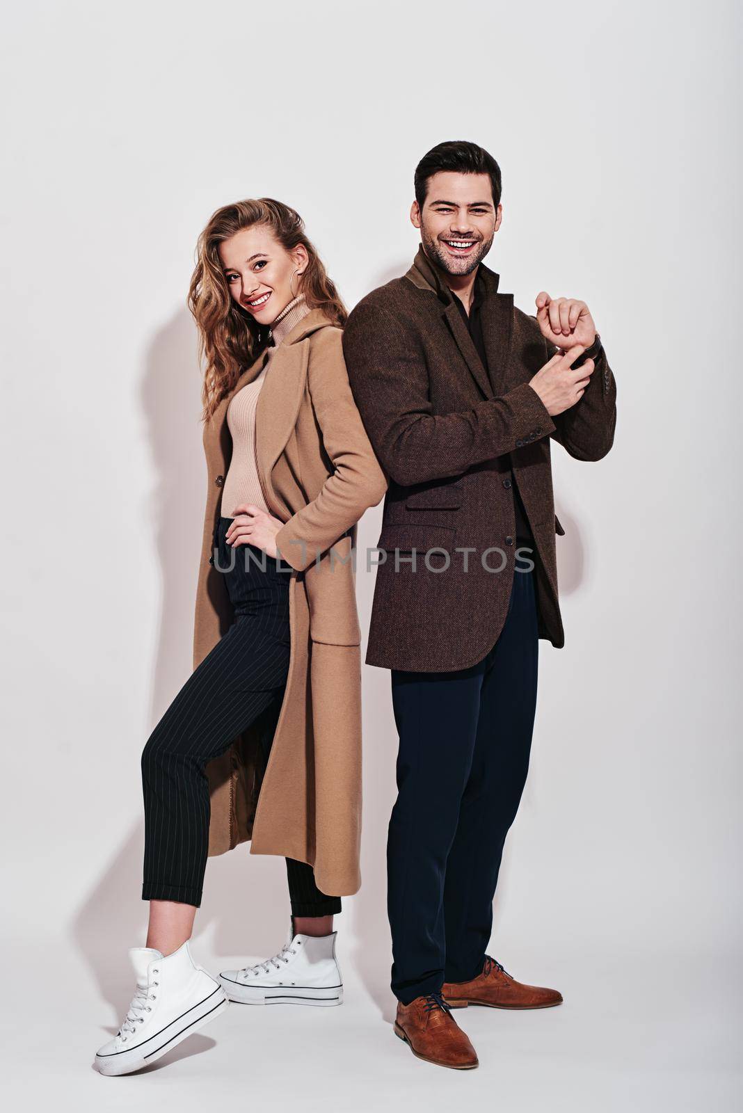 Full-length portrait of couple. Woman in beige coat, blue trousers and white converse and man wearing blue classical trousers with brown jacket. Isolated over grey background. Couple goals