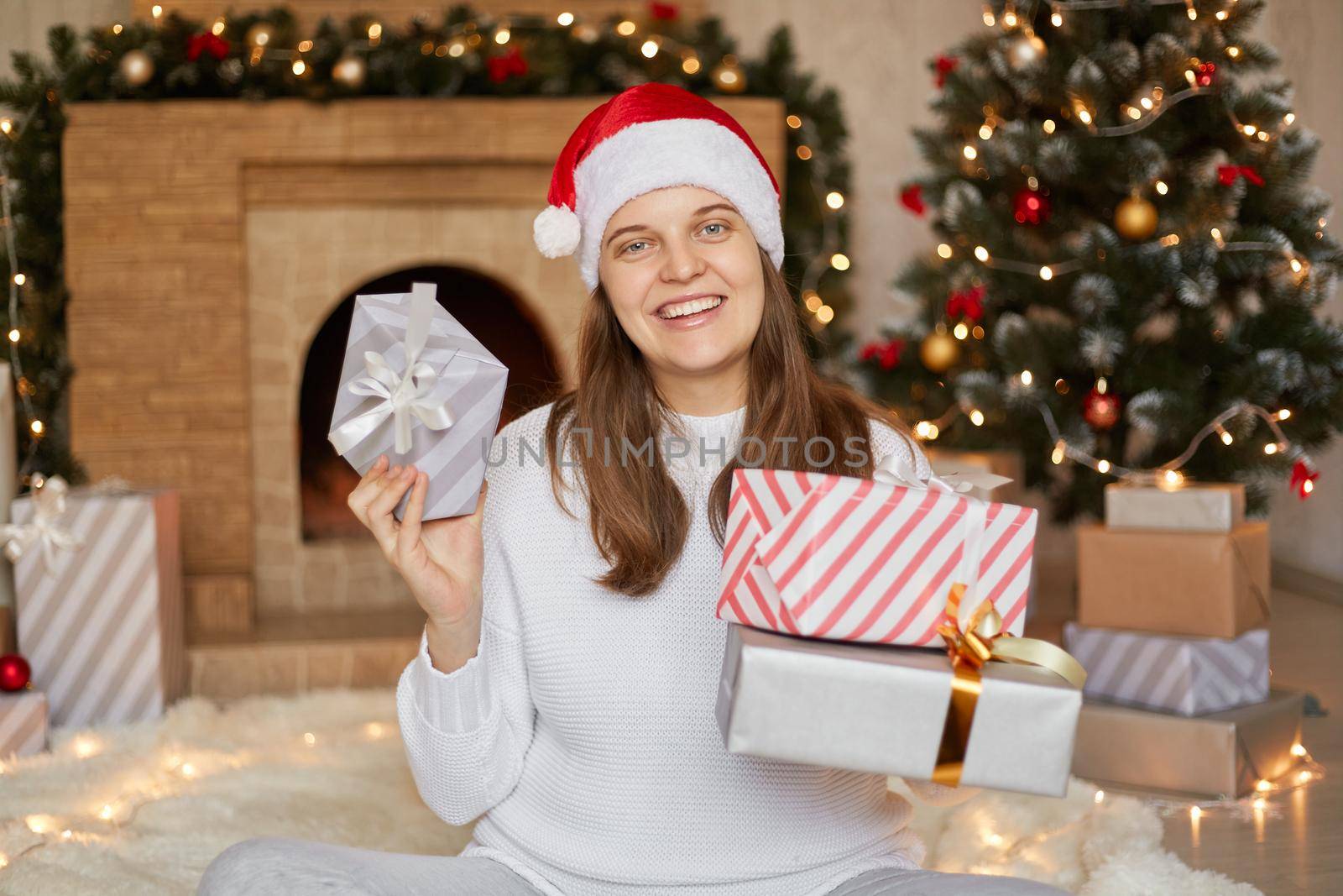 Happy excited woman in santa claus hat and white sweater with gift boxes in hands, looking directly at camera, being happy to get present for Christmas, sitting on floor on carpet near x-mas tree. by sementsovalesia