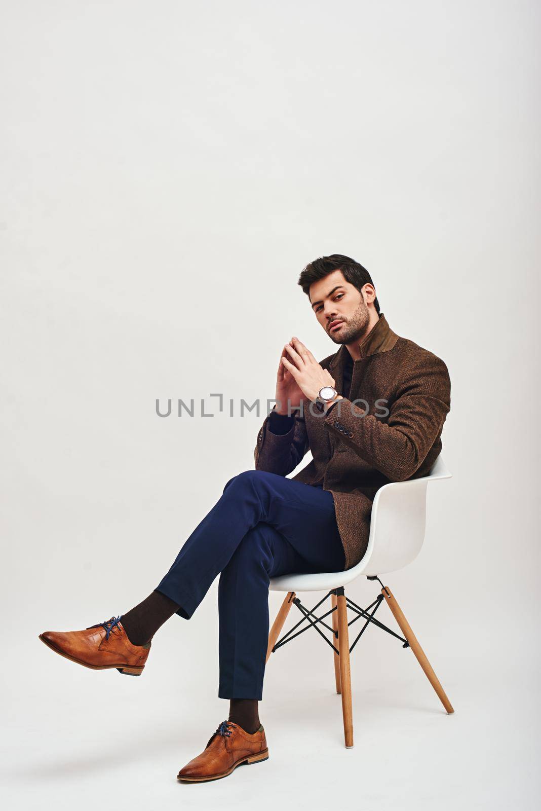 Full-length portrait of stylish dark-haired man wearing blue trousers and brown jacket, sitting on a white chair, folding his hands and looking at camera