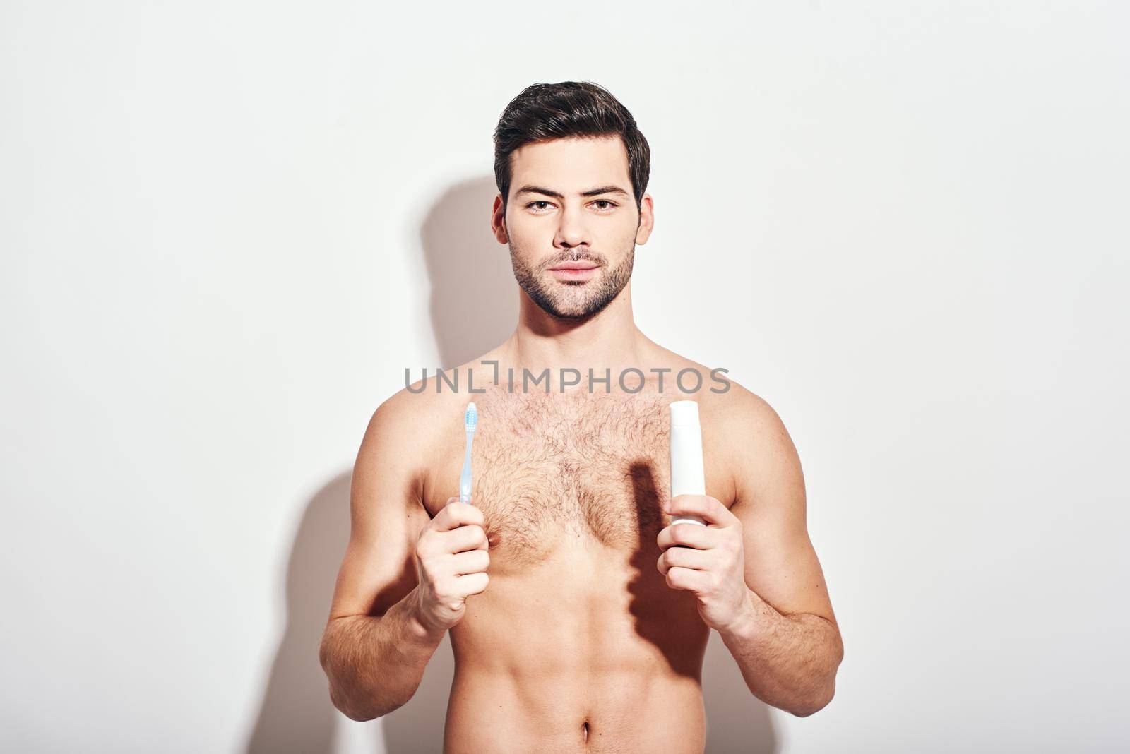 Take care of your teeth. Handsome dark-haired man holding toothpaste and toothbrush, standing shirtless isolated over white background, looking at camera by friendsstock