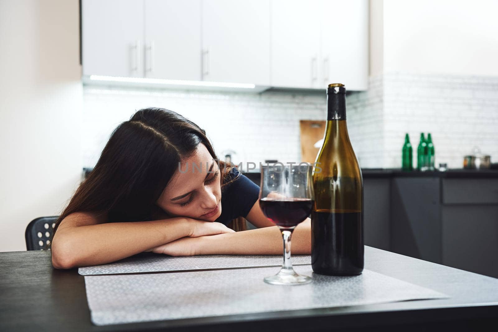 After drinking. Sleepy drunk young woman lying on the table in the kitchen and sleeping with bottle and glass of red wine standing on the table in front of her