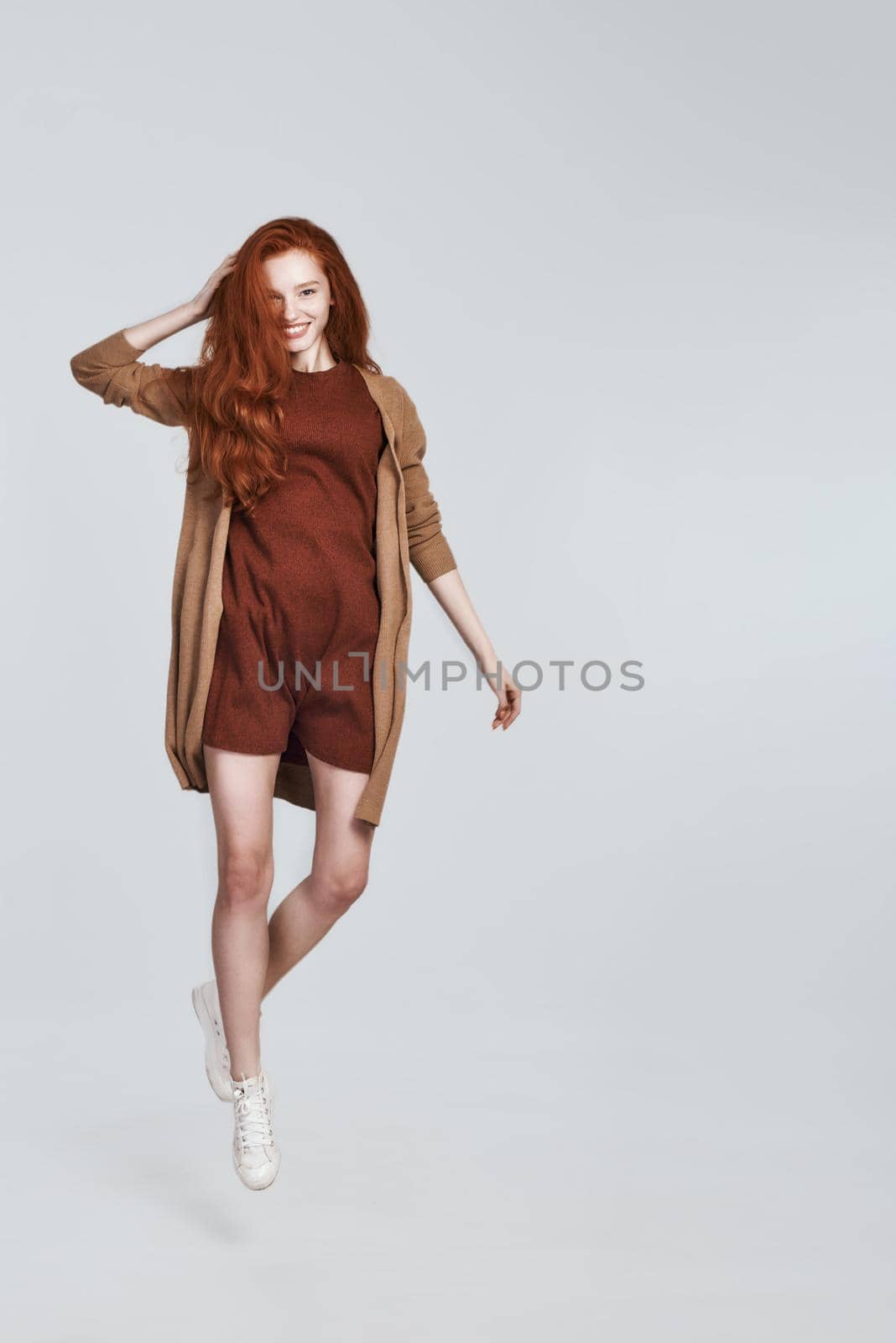 Studio shot of playful young woman with tousled red hair smiling while jumping against grey background. Beauty concept. Happiness
