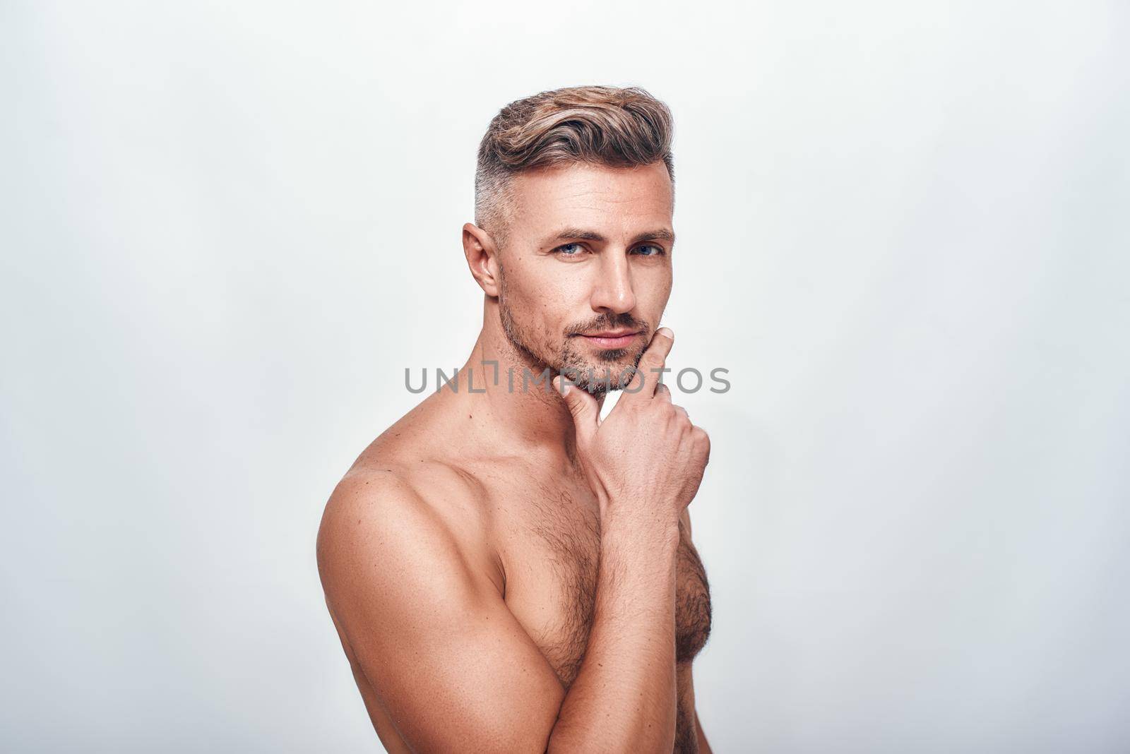 Skin care. Handsome and sexy man with a stubble keeping hand on chin and looking at camera while standing against grey background. Men beauty. Human face. Human body
