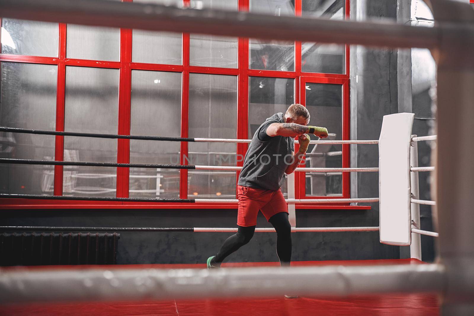 Handsome muscular sportsman in sports clothing training on boxing ring. Strong man doing shadow-boxing on red boxing ring in the gym
