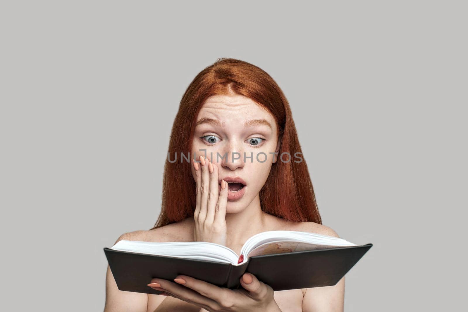 Studio shot of shocked young redhead woman reading a book and keeping mouth opened while standing against grey background