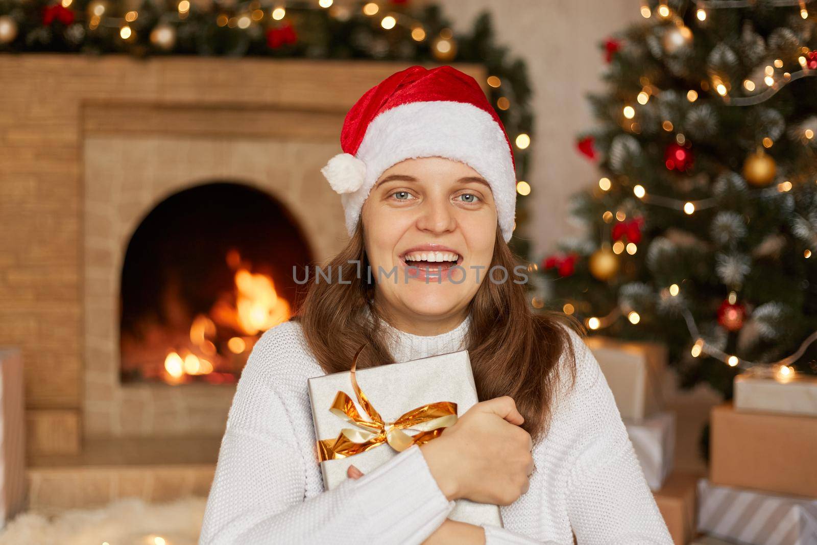 Excited woman with dark hair holding gift box in hands, has Christmas present, looks at camera with toothy smile, wearing santa hat and white sweater, fireplace and x-mas tree on background. by sementsovalesia