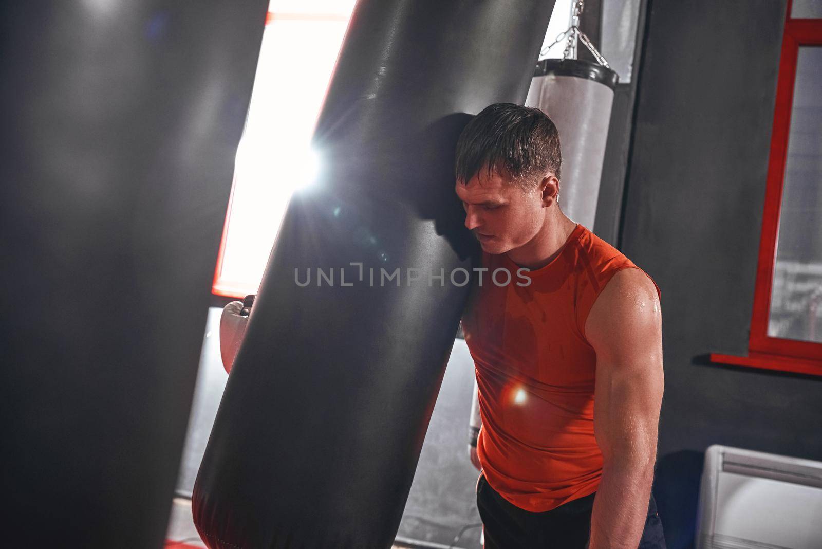 Tired after workout. Tired young athlete in sports clothing looking away after hard training on heavy punch bag in boxing gym by friendsstock