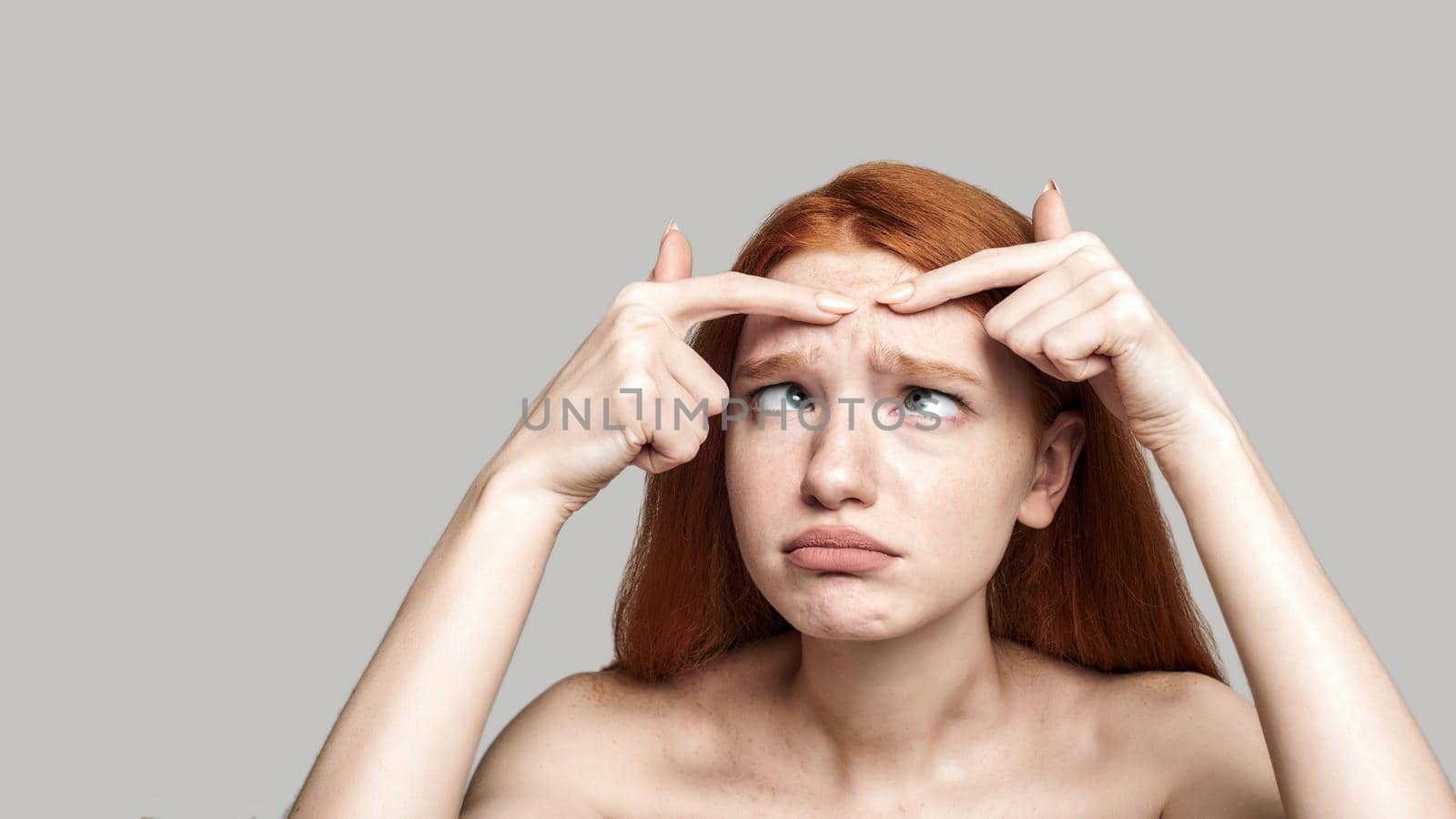 Studio shot of worried young redhead woman examining her face while standing against grey background. Skin care