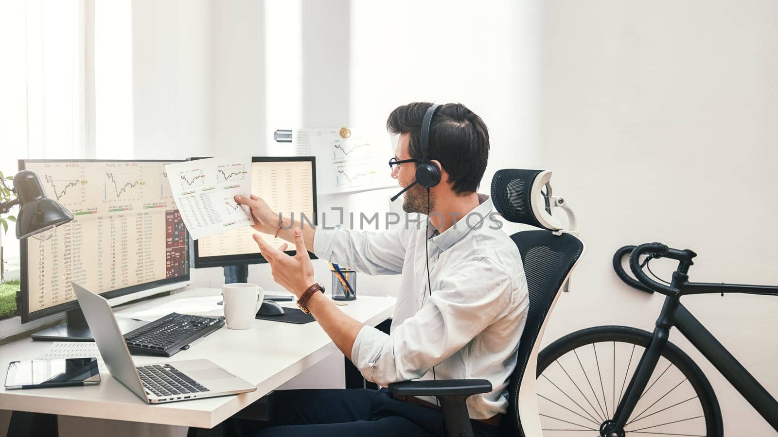 Growing chart. Side view of happy young bearded trader in headset holding financial report, talking with client and smiling while sitting in front of monitor screens in the office. Business concept. Trade concept. Call center