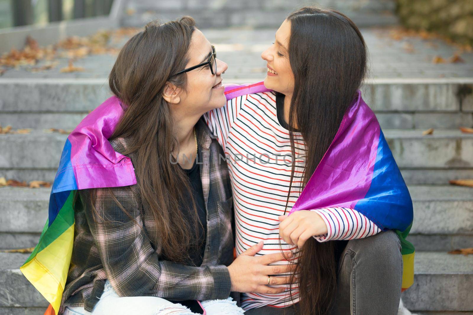Pregnant Lesbian couple, sitting on stairs hugging with rainbow flag at urban scenery. High quality photo.