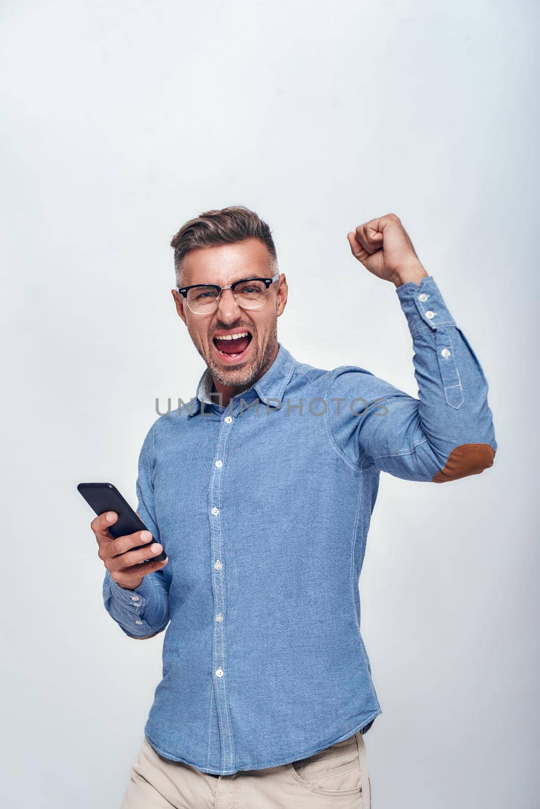 Success Portrait of handsome and happy bearded man in eyewear holding smart phone and keeping one hand clenched in fist while standing against grey background by friendsstock