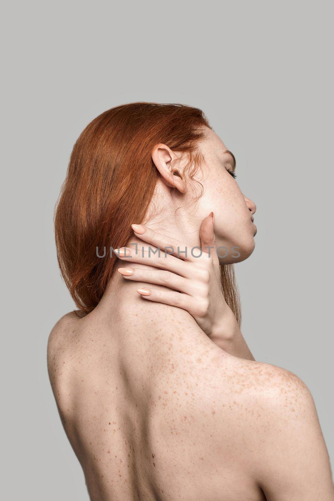 Studio shot of back view of young and beautiful redhead woman touching her neck while standing against grey background