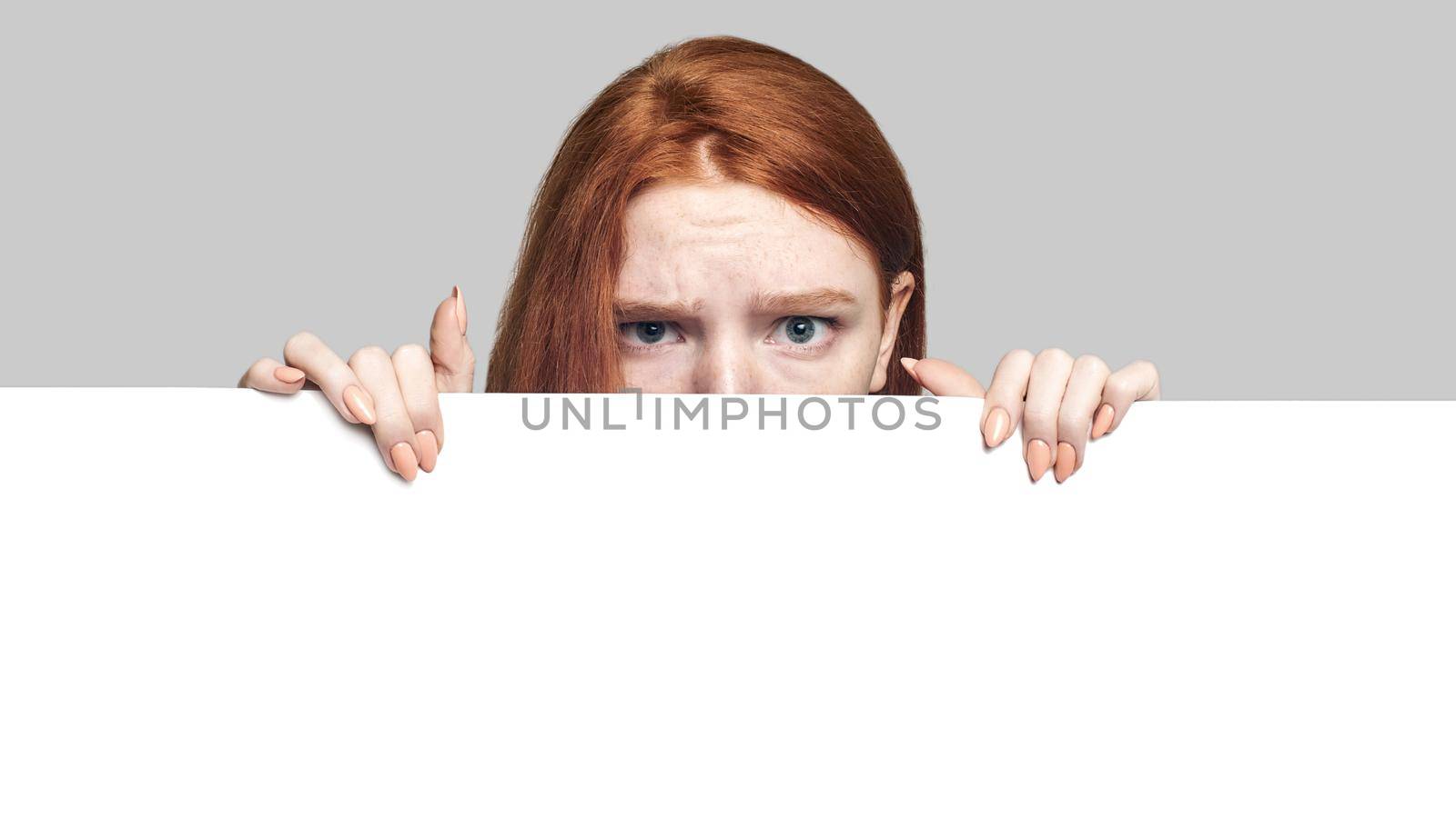 Studio shot of terrified young redhead girl looking behind empty blank board and looking at camera while standing against grey background. Advertising