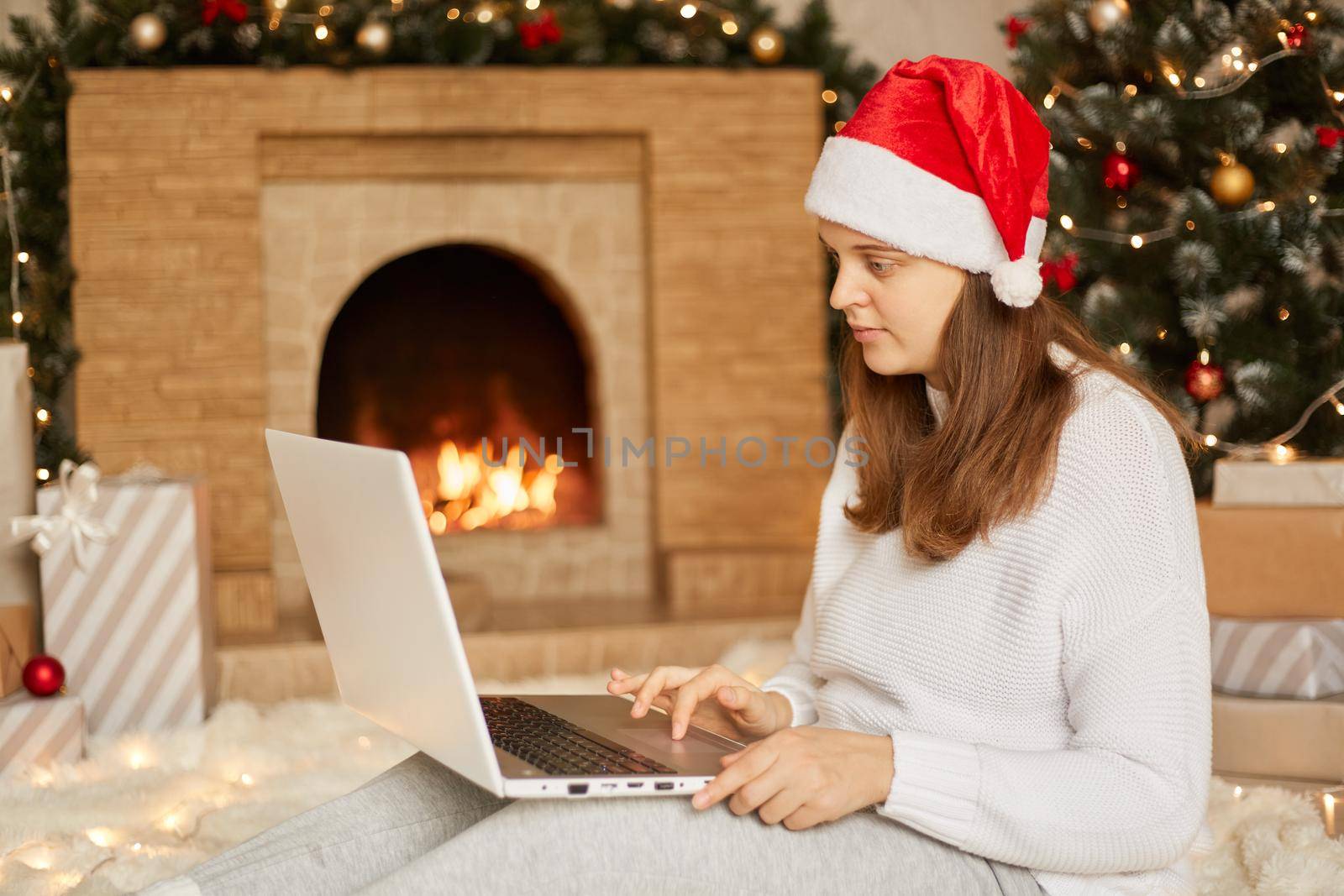 Christmas online, working via Internet. Caucasian girl at home using laptop computer notebook for video call, wears Santa hat and white sweater. Woman looking to camera of laptop, sits near fireplace by sementsovalesia