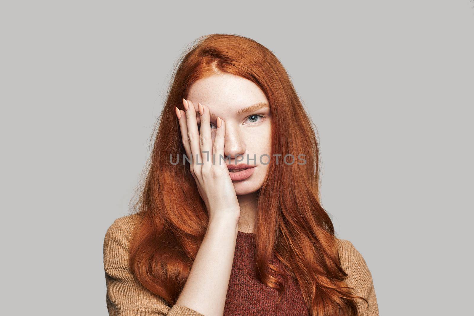 What is there Portrait of young and cute teenage girl with red silky hair covering face with hand and looking at camera while standing against grey background by friendsstock
