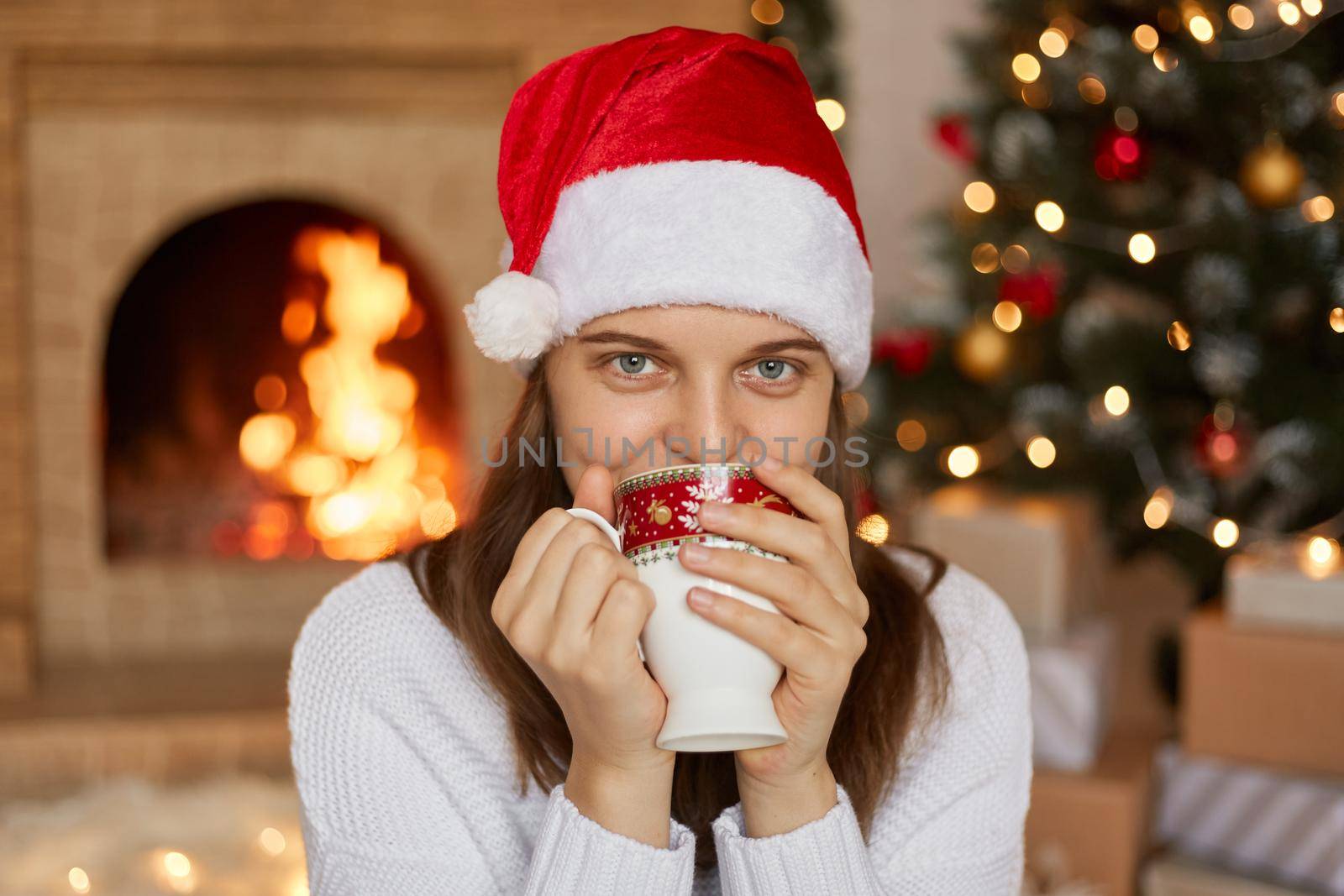 Beautiful young woman wearing Santa Claus red hat sitting near fire place in living room with x-mas decoration, lady drinking hot beverage, holding cup with both hands, looks at camera. by sementsovalesia