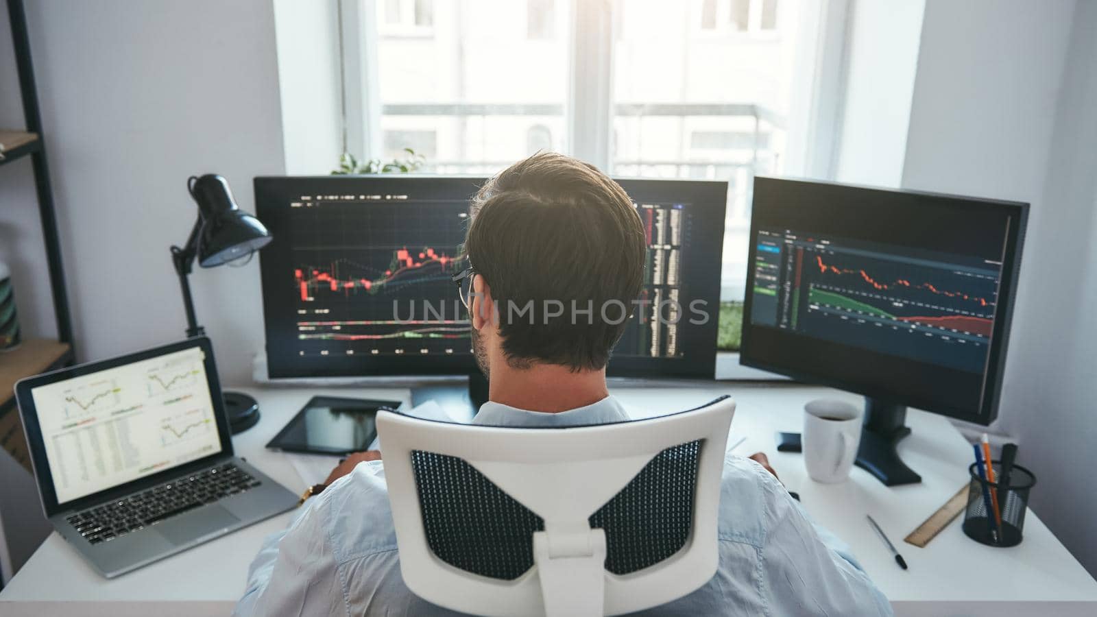 Trading stocks online. Back view of young businessman or trader working with graph and charts on computers at his modern office by friendsstock