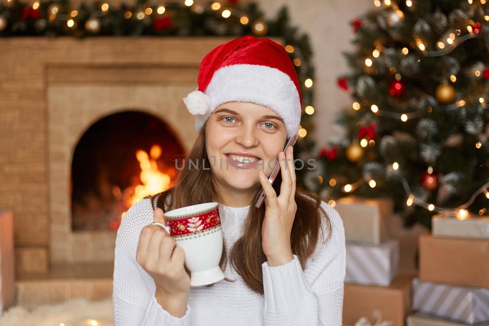Beautiful woman with christmas hat drinking hot beverage while talking phone with somebody, greets with New year, expresses happiness, wearing white jumper, posing indoor near fireplace and x-mas tree