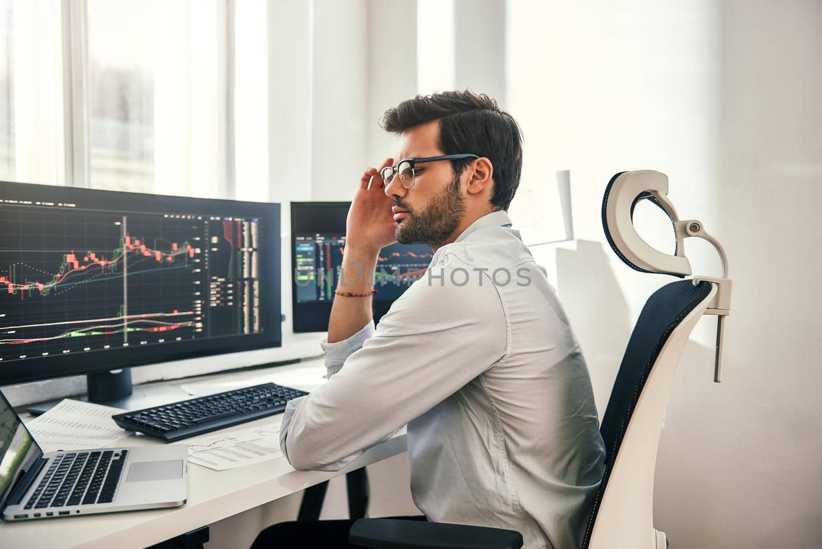 Full concentration. Young and smart businessman in formal wear is adjusting his eyeglasses and using laptop while while sitting in his office in front of computer screens with trading charts. Stock exchange. Trade concept. Investment concept