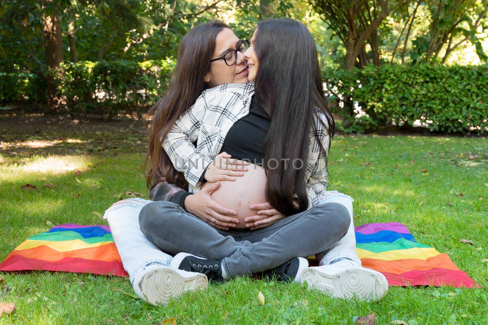 Portrait of affectionate pregnant lesbian couple with rainbow flag, relaxed at the park. Two happy girlfriends. Free same-sex love. Homosexual relationship. LGBT Community Pride by HERRAEZ