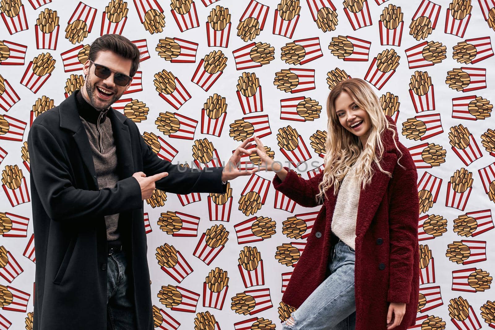 We love french fries. Young couple in eco fur coats posing at the street brand wall background. Brand wall designed with street food. Cold season. Young man in sunglasses huggs his blond haired girlfriend.