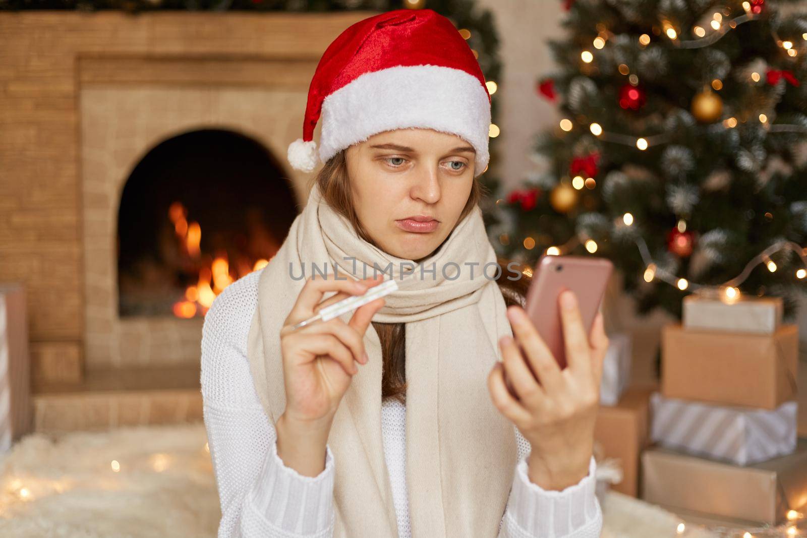 Young woman wearing Christmas hat, take video calls during corona virus pandemic, with Christmas tree and fireplace on background, showing thermometer to camera, telling about her high temperature. by sementsovalesia