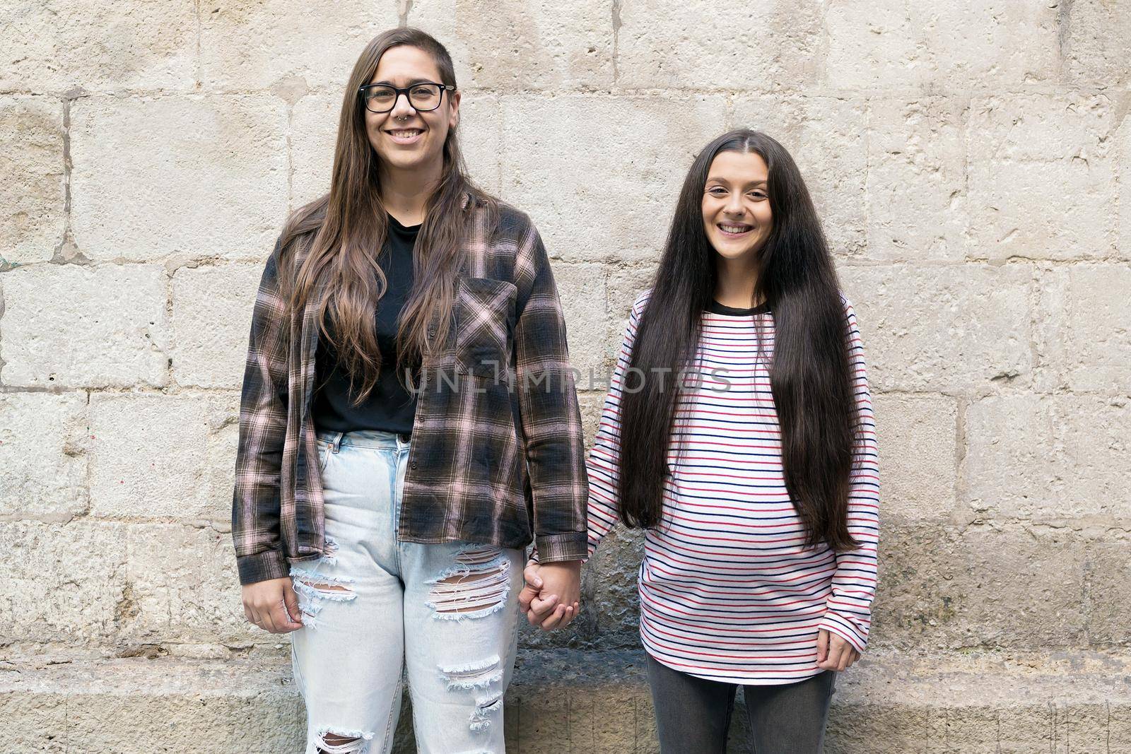 Two young lesbians holding each other and standing near a stone wall, smiling, happy. High quality photo