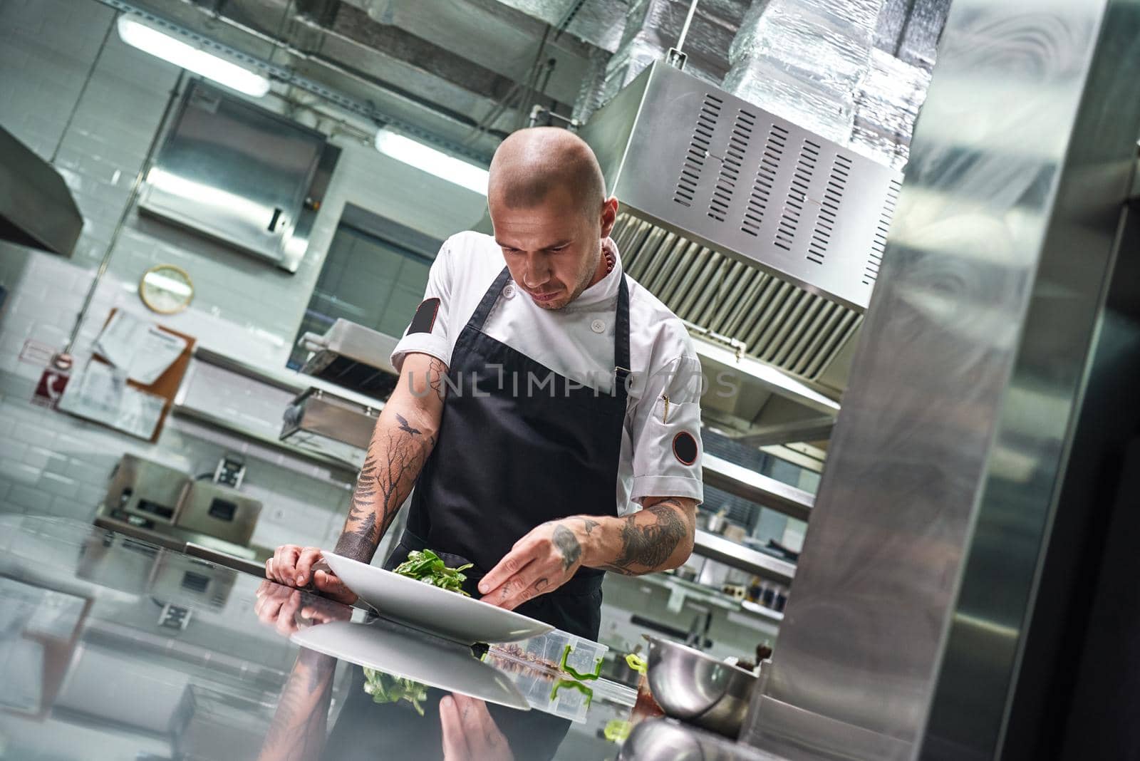 Food concept. Portrait of handsome professional chef in black apron decorating a salad on the plate while working in restaurant kitchen. Ready to serve