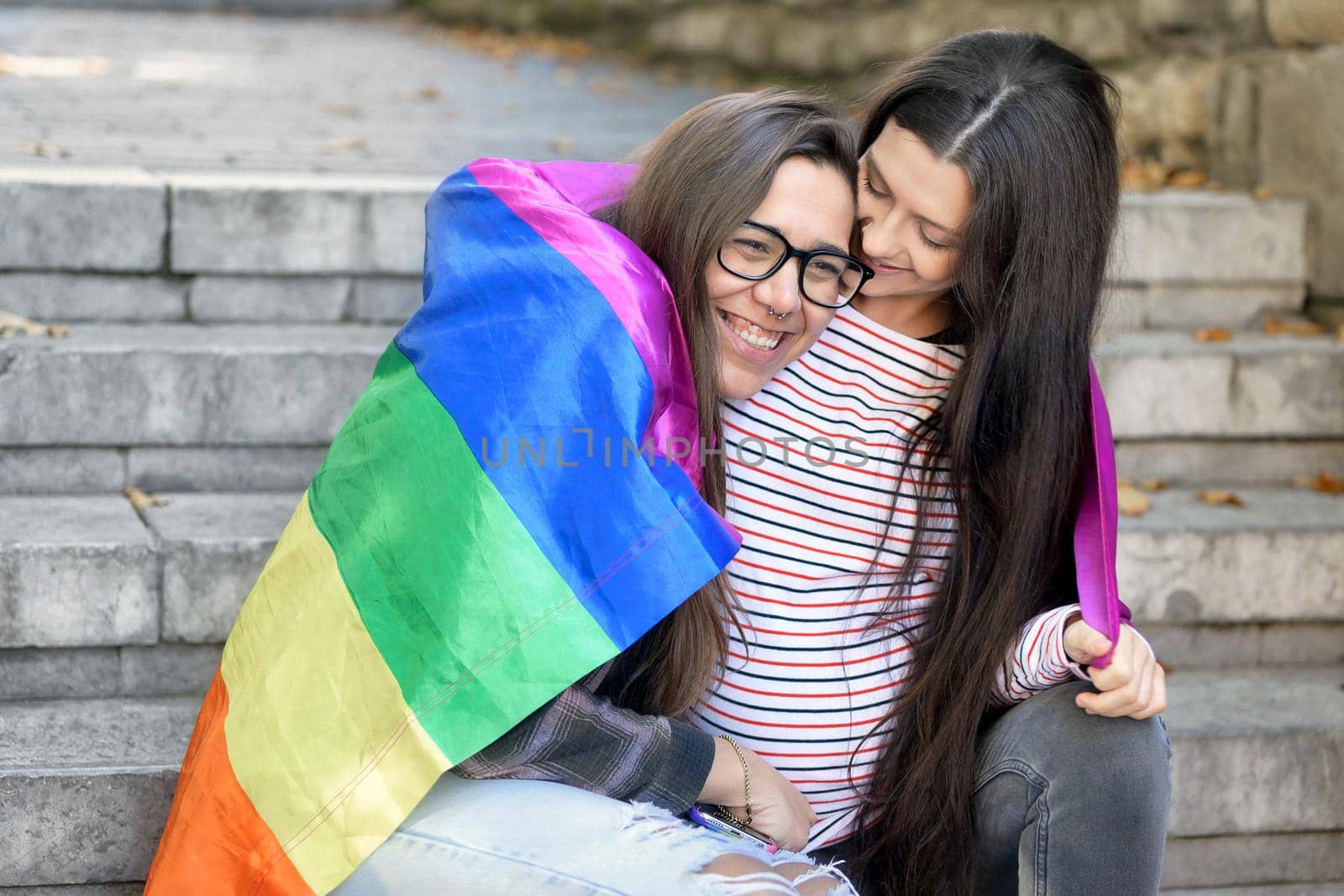 Lesbian couple sitting on steps hugging with rainbow flag at urban scenery. High quality photo.