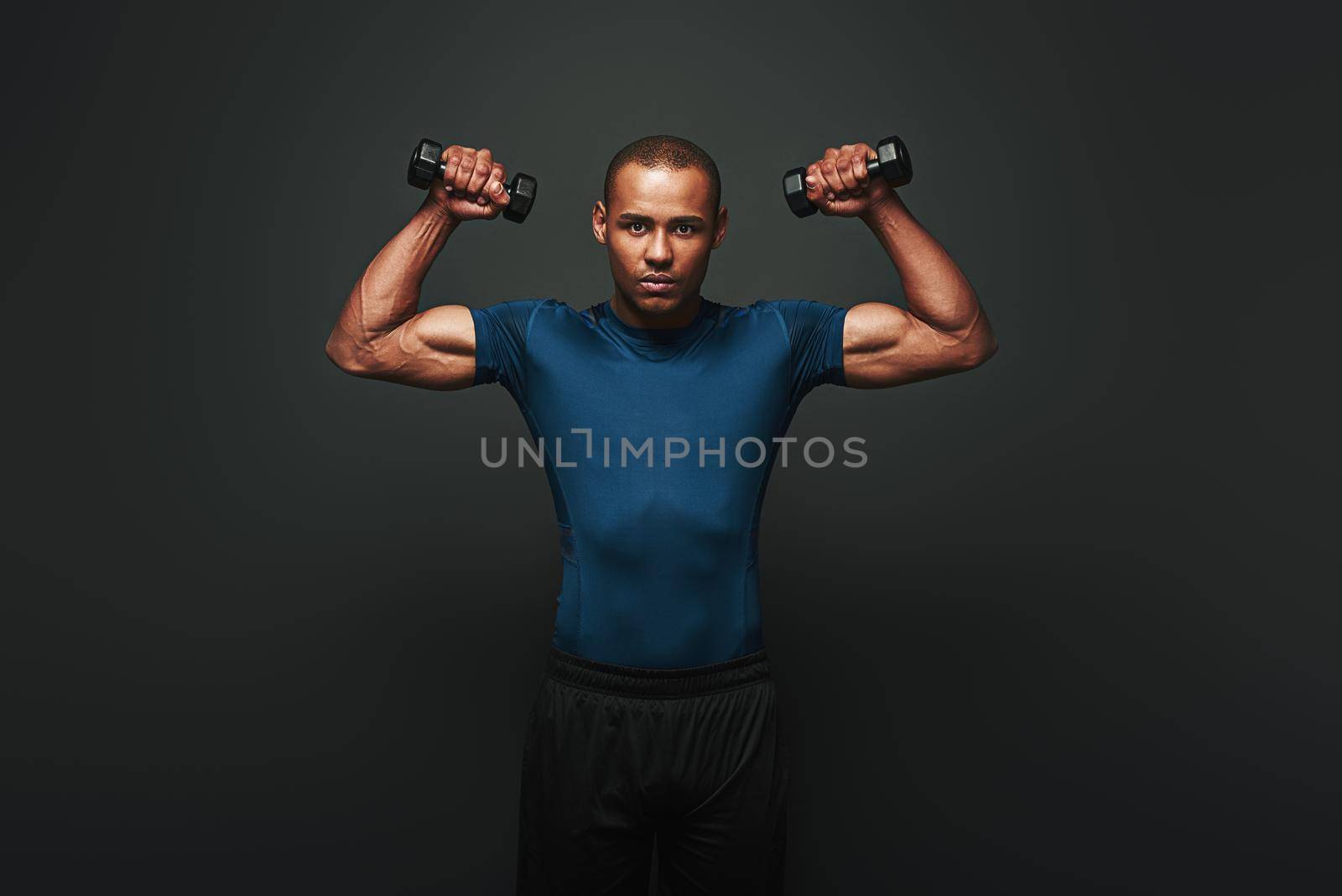 Athletic young sportsman exercising with dumbbells and looking at camera isolated on dark background. He demonstrates his perfect muscles