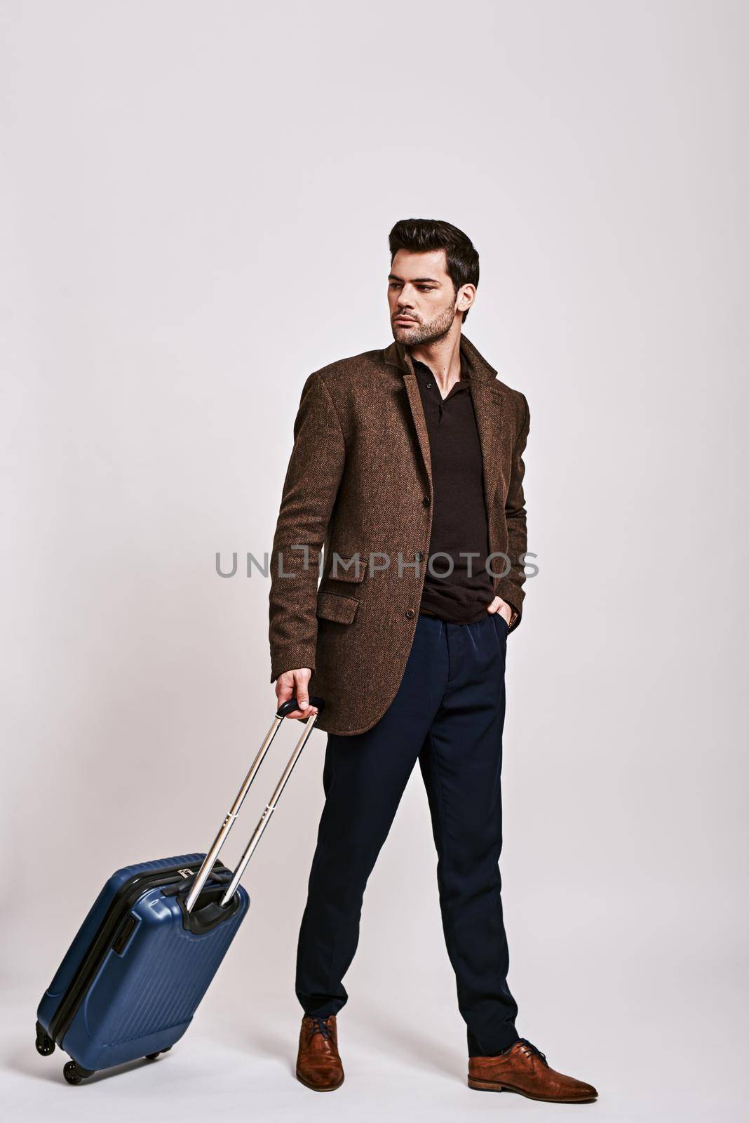 Full-length portrait of stylish dark-haired man wearing blue trousers, brown jacket standing with suitcase and looking away isolated over grey background
